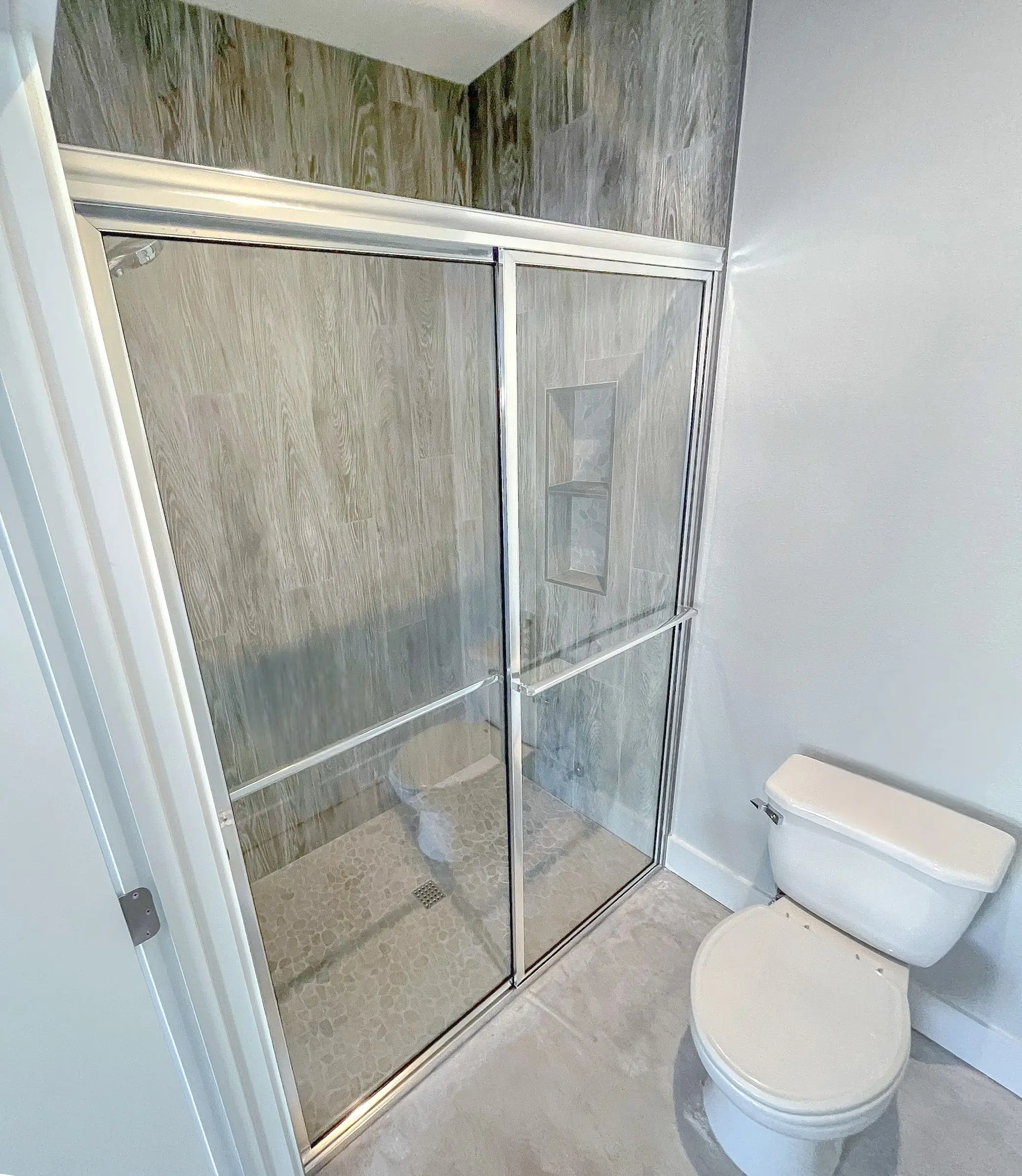 Chrome-framed bypass sliding shower doors in a compact bathroom, with wood-look porcelain tile and a pebble mosaic floor.