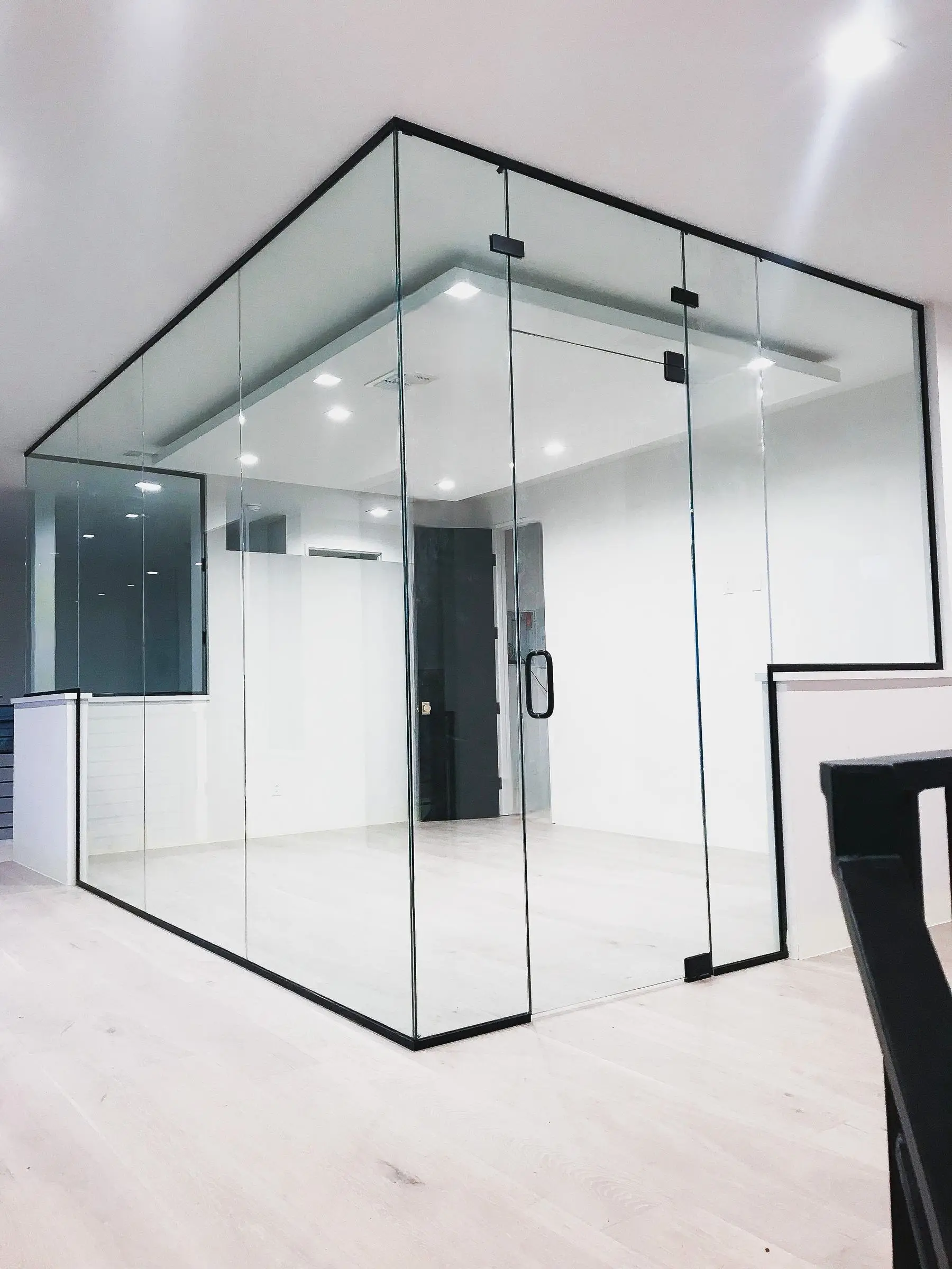 A frameless glass office enclosure with slim black framing at the floor and ceiling, a glass swing door with a black pull handle, and pale wood flooring.