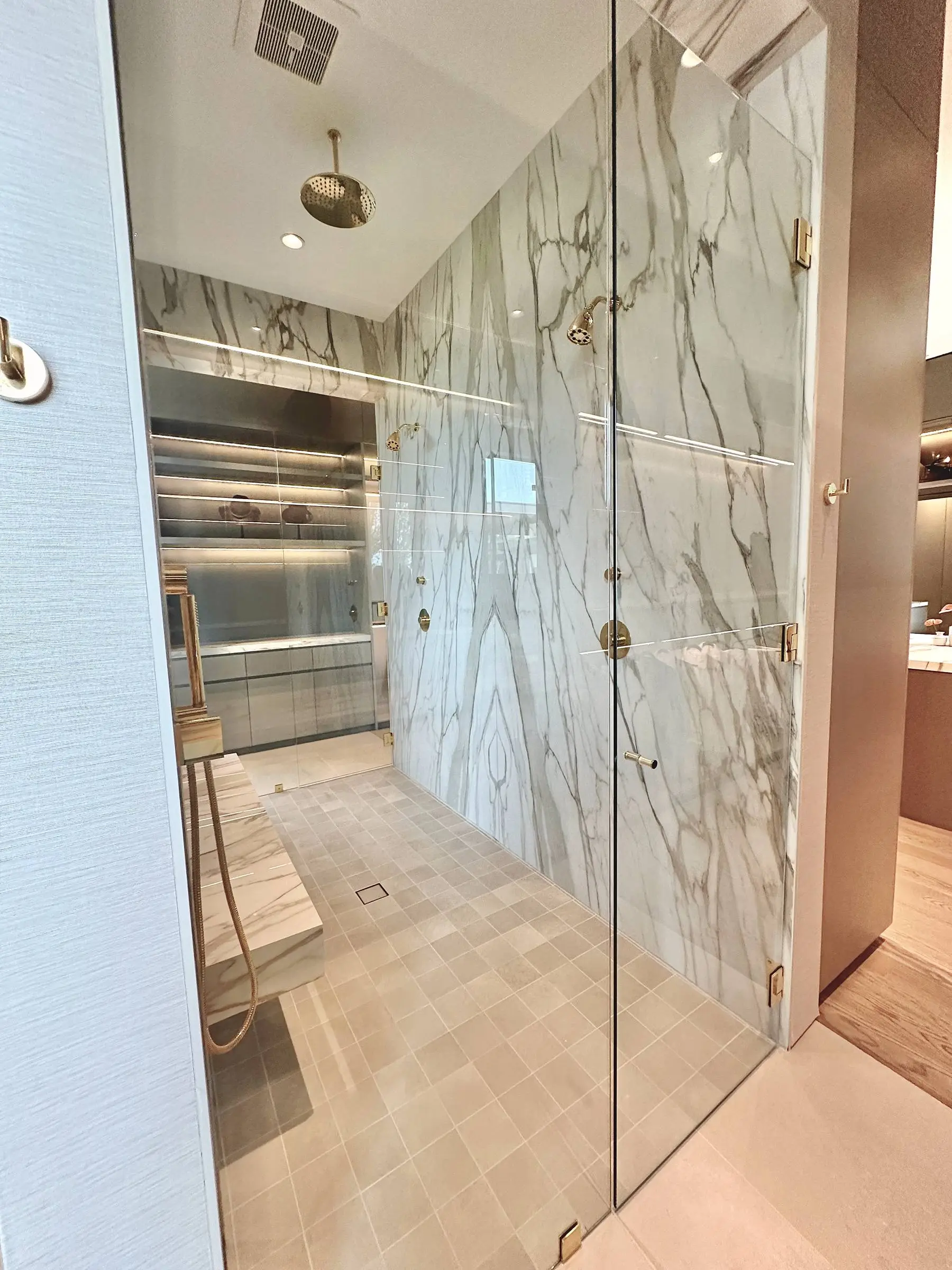 A frameless glass shower with book-matched marble slab walls, a marble bench, brass fixtures, and a clear glass door on brass hinges.