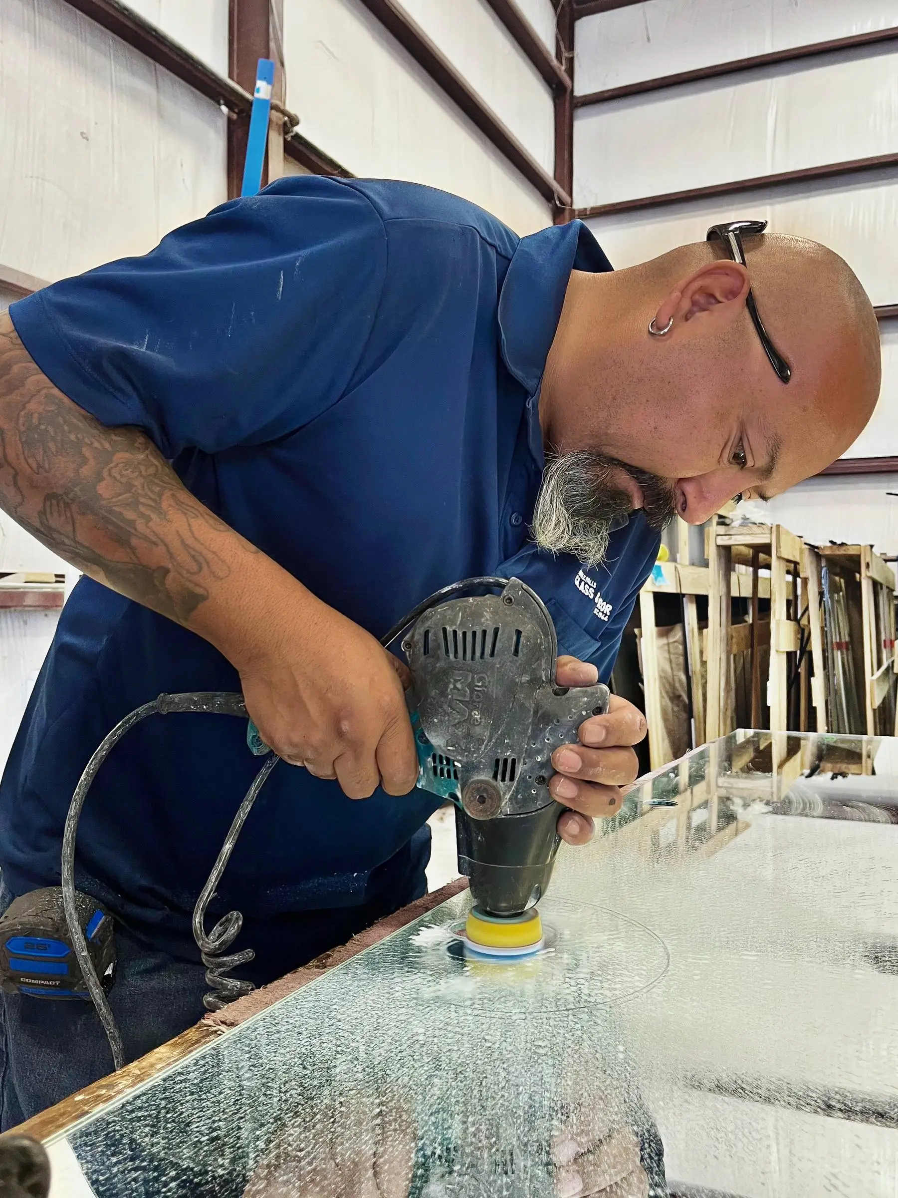 A Marble Falls Glass shop hand in a company polo running a wet grinder along the edge of a glass panel, with racked glass behind him.