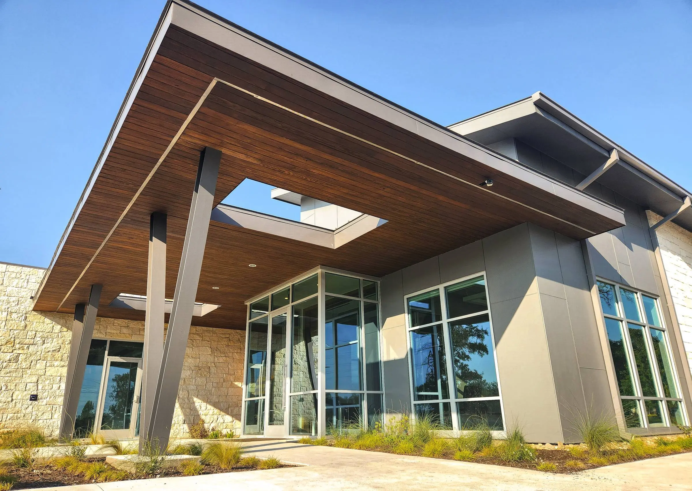 A Central Texas commercial building with a limestone base, metal panel walls, a deep wood soffit over the entry, and aluminum-framed glazing running the length of the front.