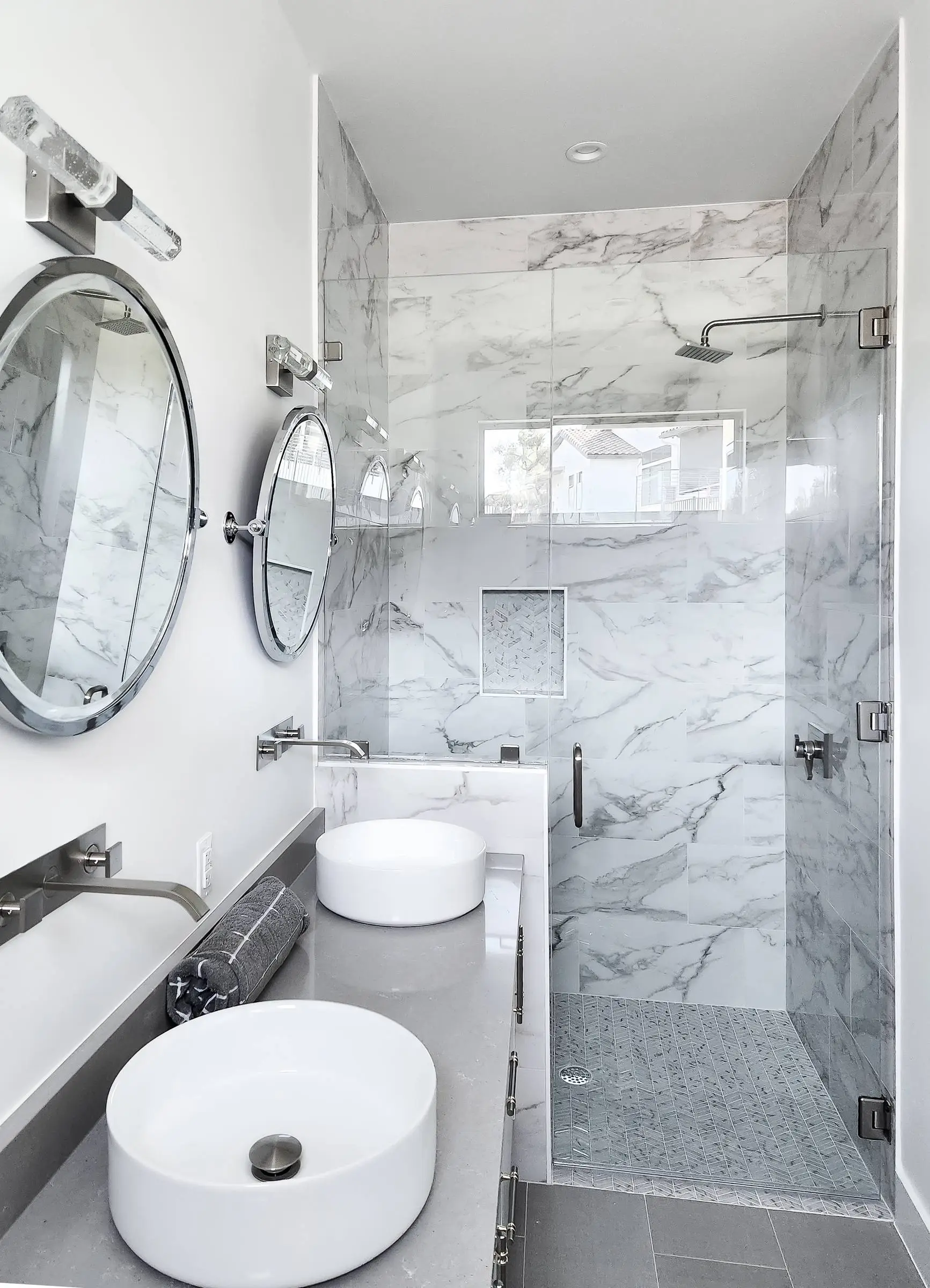 Two round chrome-framed pivot mirrors above vessel sinks, with a marble-tiled walk-in shower and frameless glass door behind them.