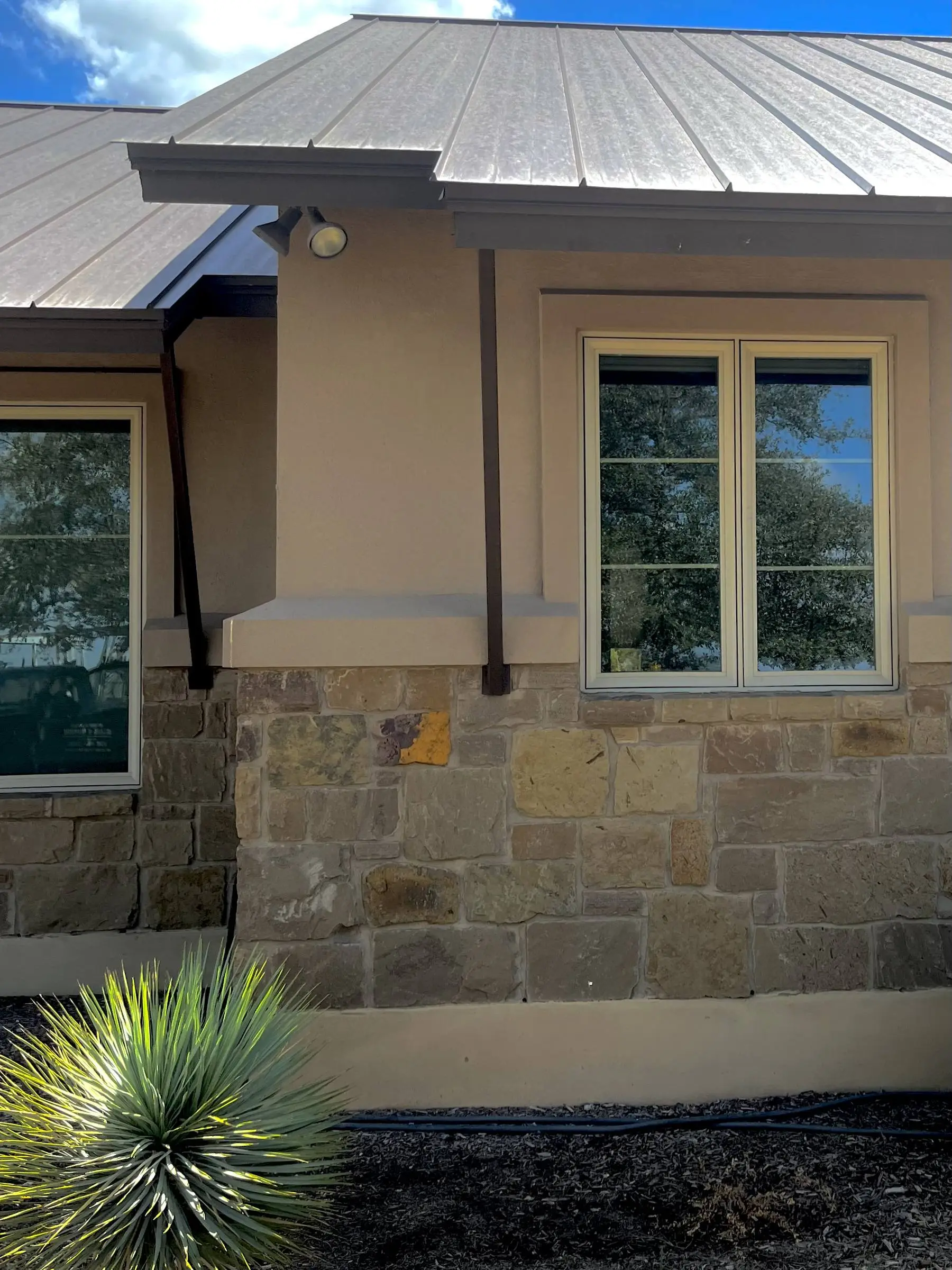 The exterior of a Hill Country home with a standing-seam metal roof, stucco walls over a limestone base, and two tan-framed windows looking onto live oaks.